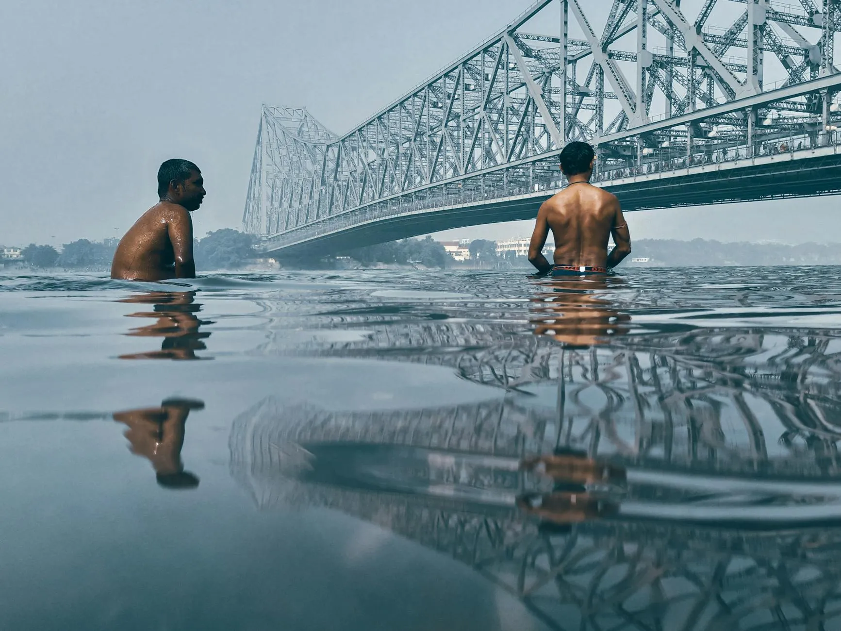 Kolkata Airport (CCU) — Men enjoy a swim in the Hooghly River with Howrah Bridge in the backdrop, Kolkata.