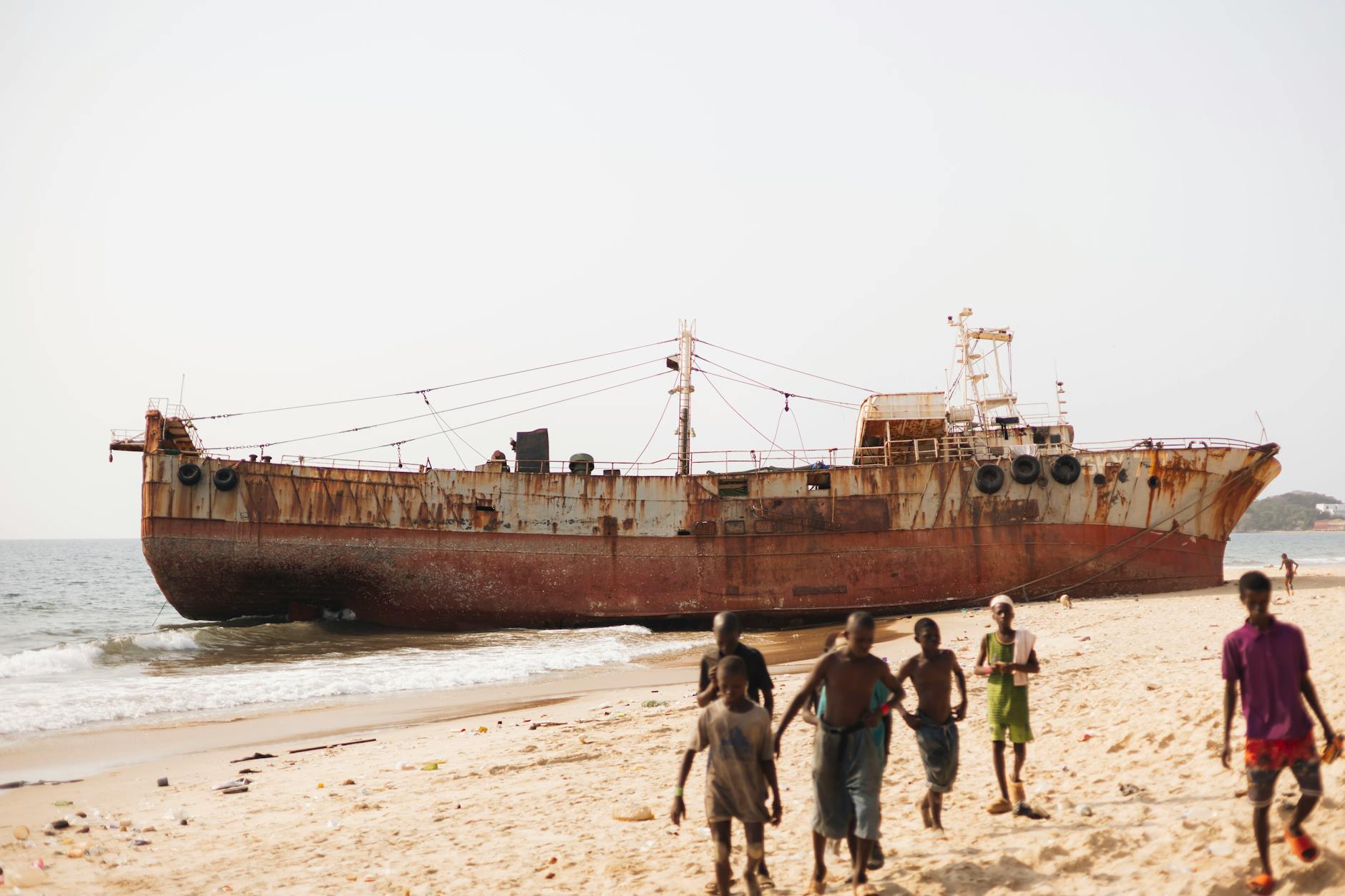 Rusty shipwreck on a sunny beach in Freetown with children playing nearby.