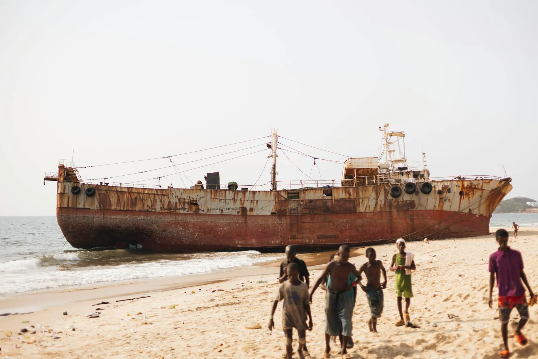 Rusty shipwreck on a sunny beach in Freetown with children playing nearby.
