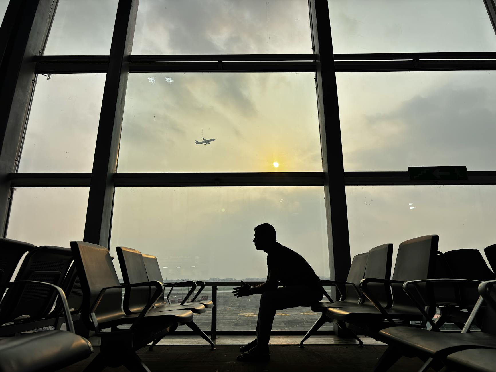 Silhouette of a man sitting at Kolkata Airport with a plane in the sky during sunset.