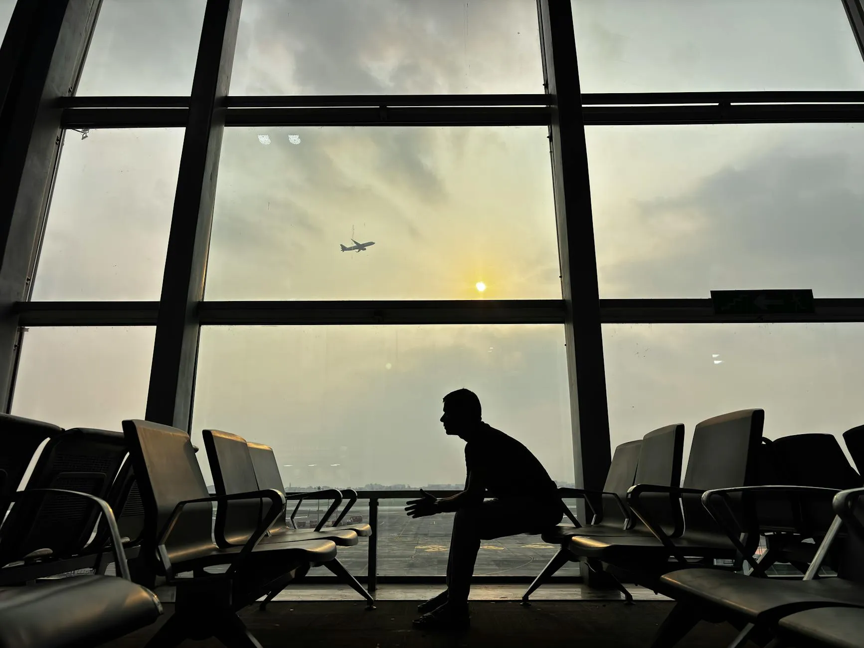 Silhouette of a man sitting at Kolkata Airport with a plane in the sky during sunset.