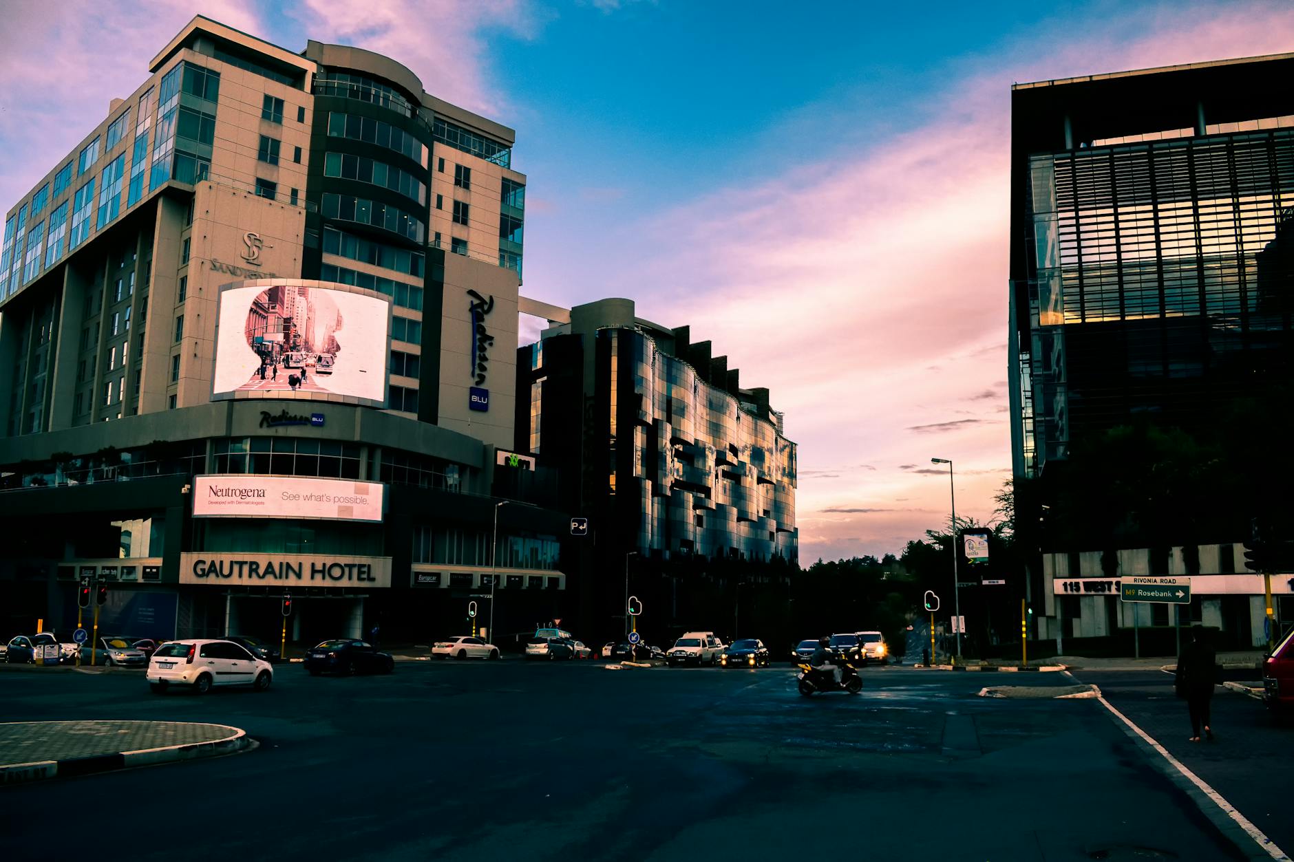 A bustling city street with modern skyscrapers and vehicles at sunset, Gautrain Hotel visible.