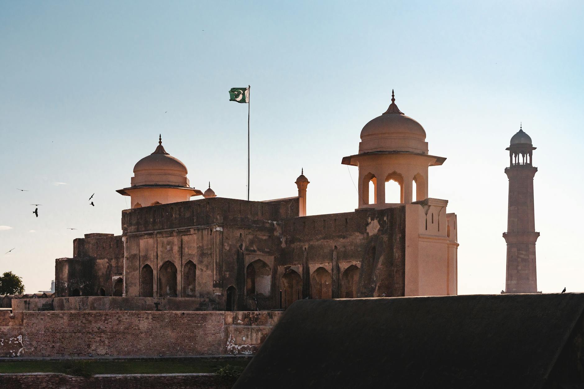 A view of Lahore Fort with a minaret silhouette against a vibrant sunset sky, featuring the Pakistani flag.