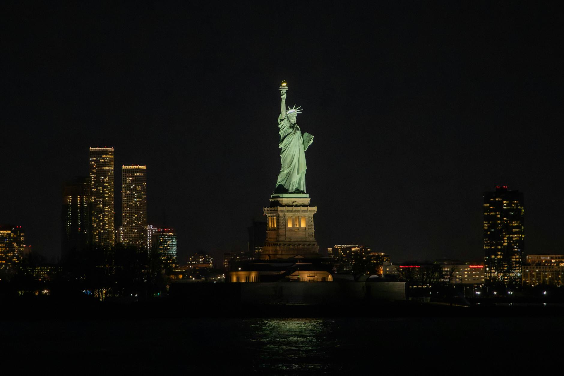 Dramatic night view of the Statue of Liberty against the New York City skyline, illuminating the harbor.