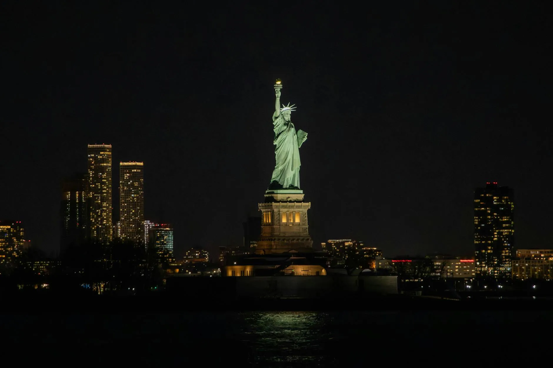 Dramatic night view of the Statue of Liberty against the New York City skyline, illuminating the harbor.