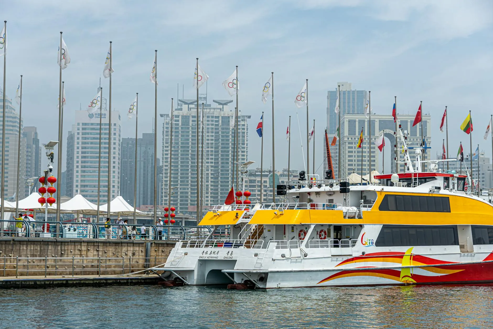 A bright yellow ferry moored in an urban harbor with skyscrapers and flags in the background.