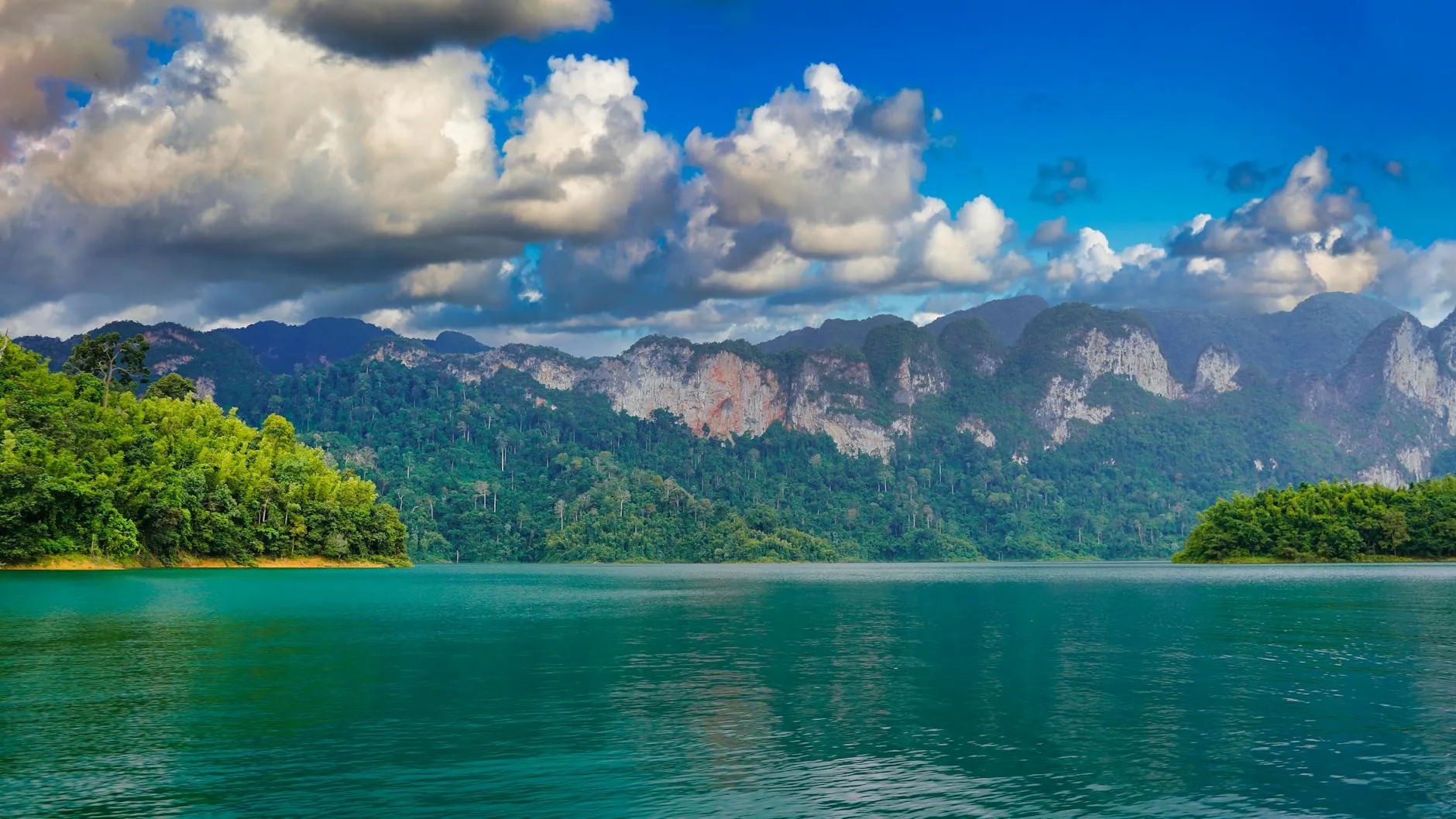 Surat Thani, Thailand — Breathtaking view of a serene lake and majestic mountains under a cloudy sky in Surat Thani, Thailand.