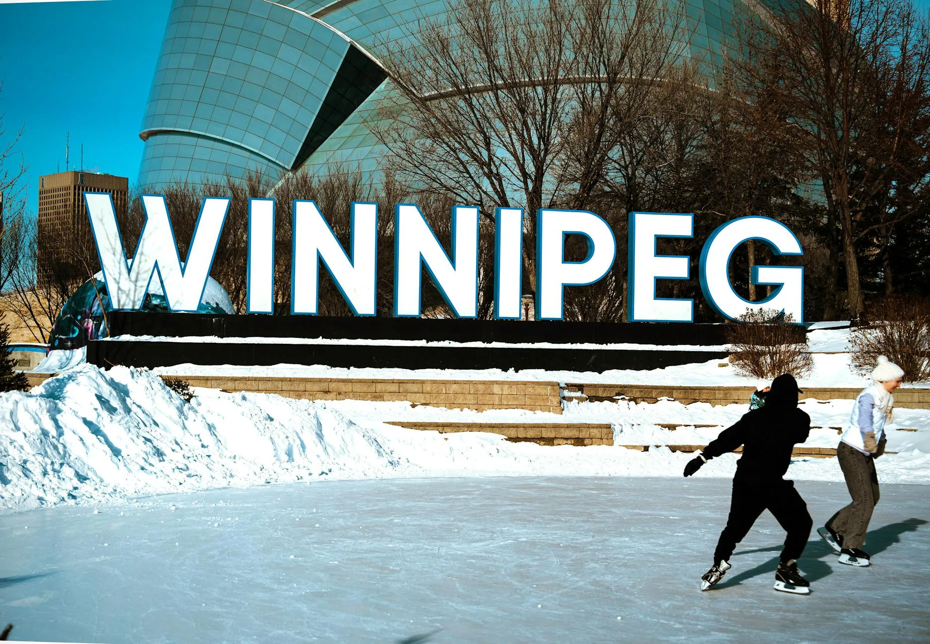 People ice skating in front of the iconic Winnipeg sign on a sunny winter day.
