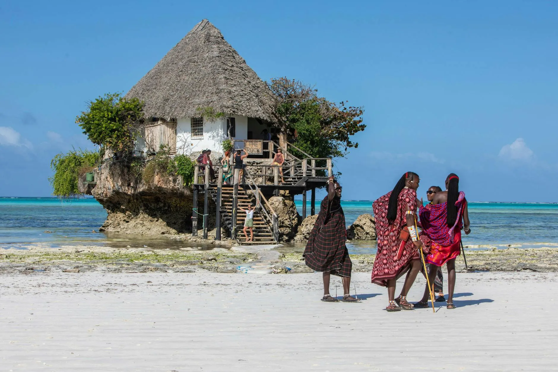 Cultural scene with The Rock restaurant and locals in traditional attire on a Zanzibar beach.