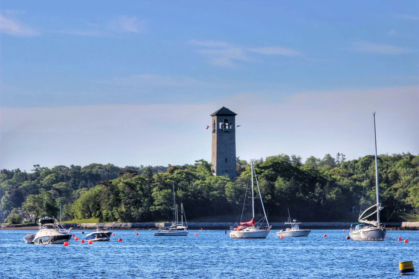 Sailboats moored in Halifax harbour, Nova Scotia, with a historic stone tower above the wooded shoreline under a clear blue sky