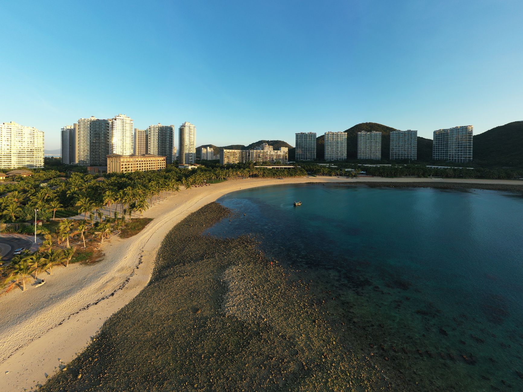 Aerial view of Sanya's beachfront with city skyline, palm trees, and clear water.