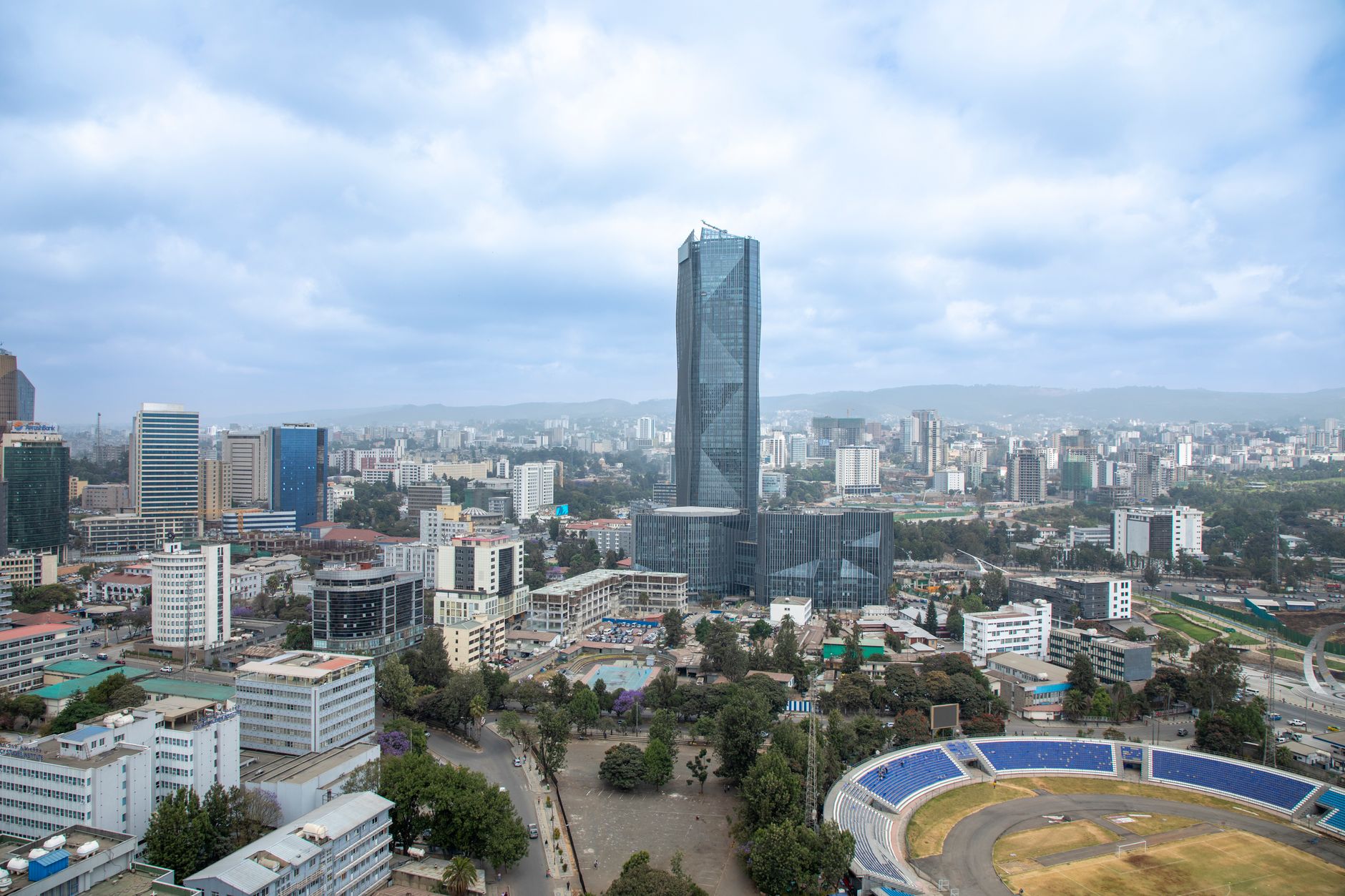 An aerial view of Addis Ababa, a tall faceted glass tower rising over the city centre with a stadium and tree-lined streets below and the surrounding hills on the horizon.