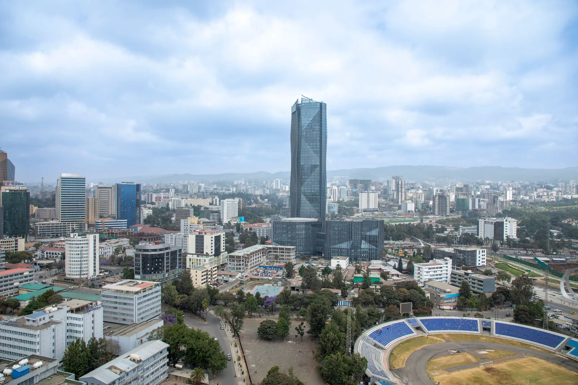 An aerial view of Addis Ababa, a tall faceted glass tower rising over the city centre with a stadium and tree-lined streets below and the surrounding hills on the horizon.