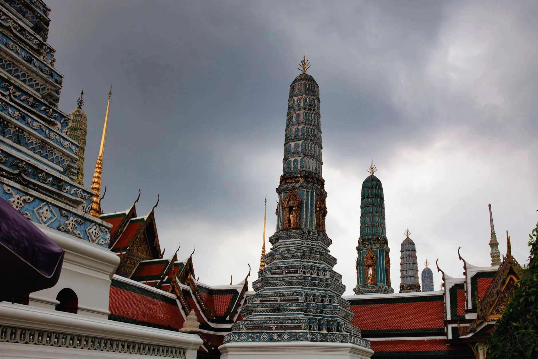 The porcelain-tiled spires and gilded roof finials of Wat Arun in Bangkok, Thailand, against a dramatic grey sky.