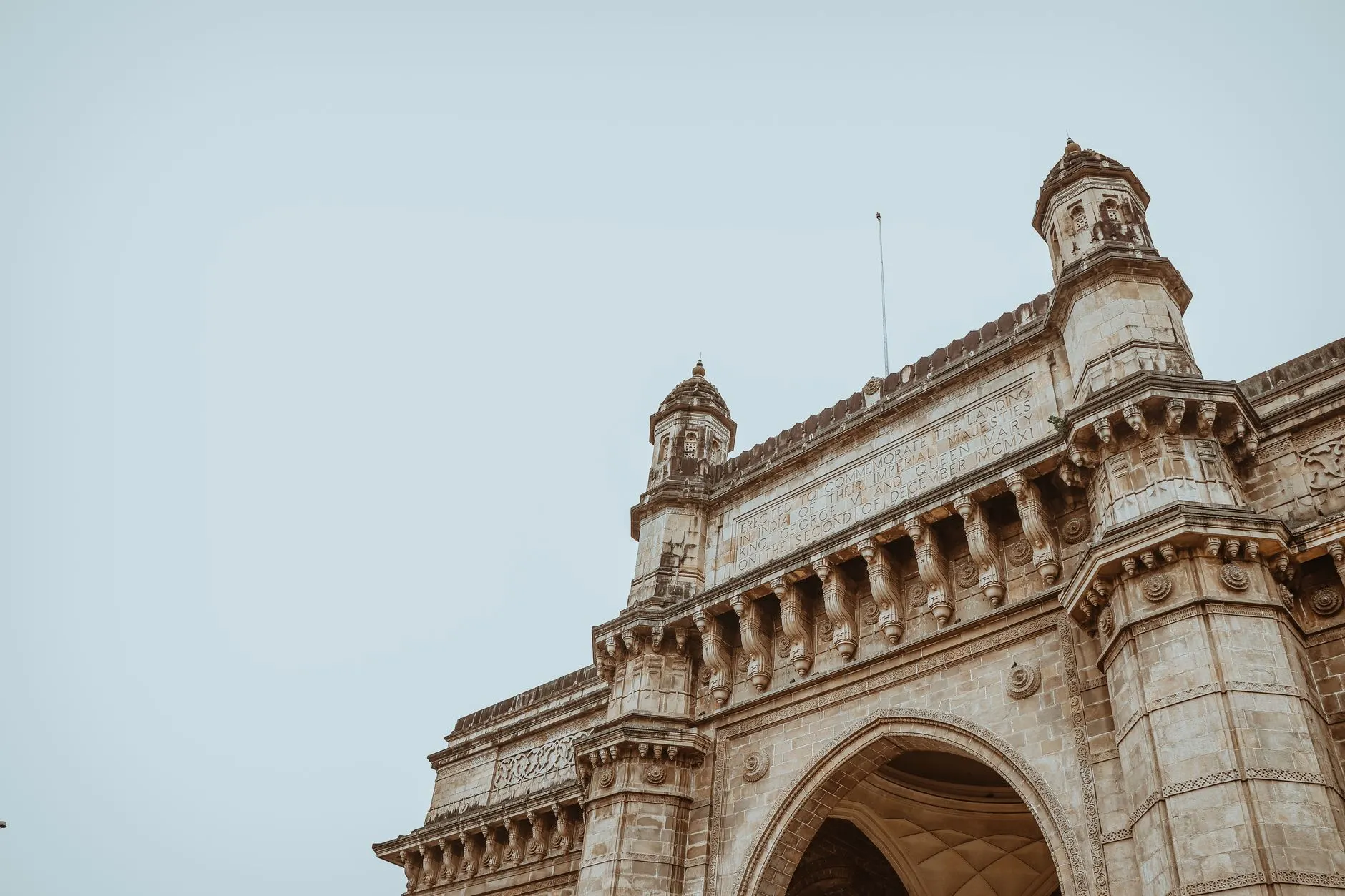 A low-angle view of the Gateway of India in Mumbai, its honey-coloured stone arch and carved turrets against a pale sky, the commemorative inscription legible above the archway.
