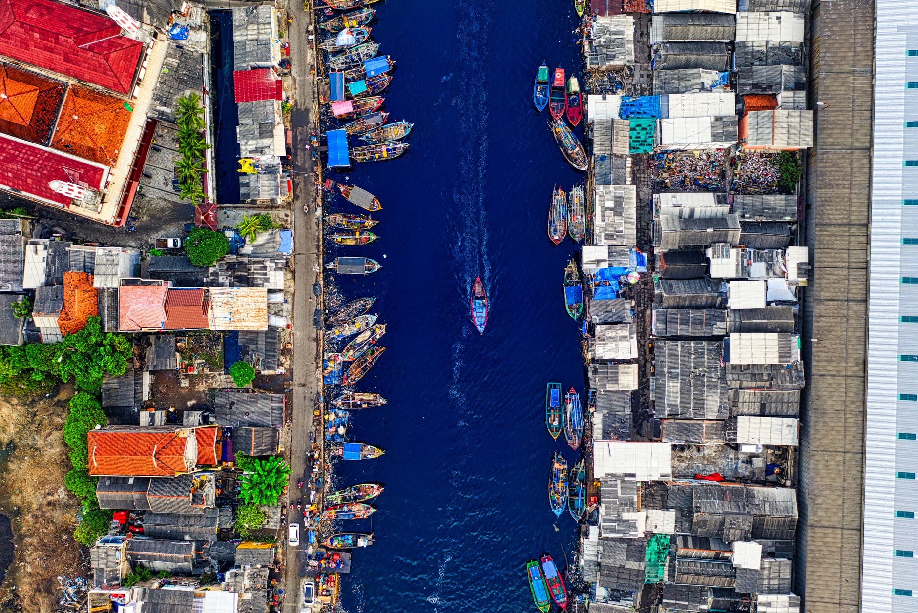An overhead view of a Jakarta harbour canal, both banks lined with brightly painted wooden fishing boats moored beside dense red- and grey-roofed houses, with one boat under way down the middle.