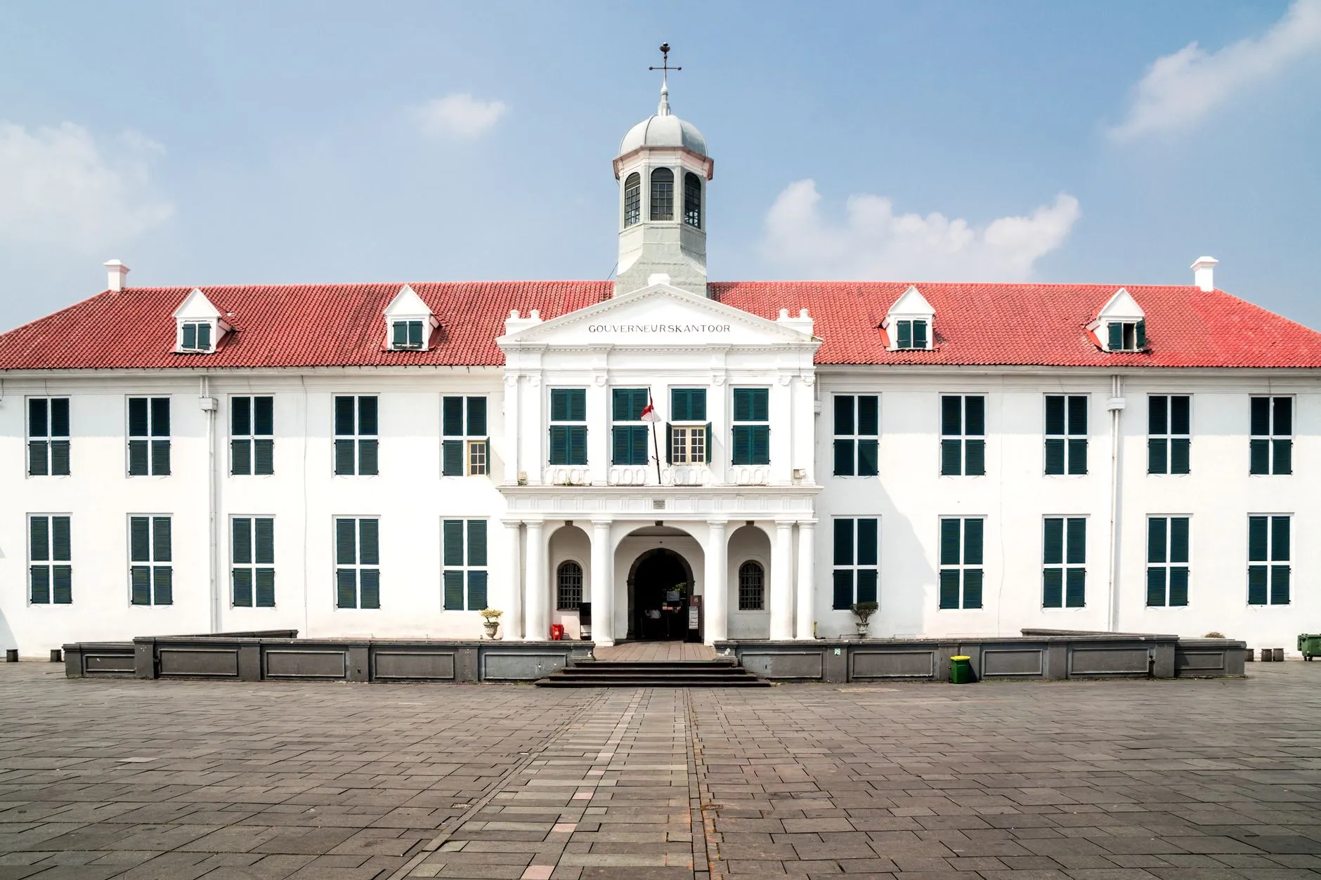 The white colonial facade and red-tiled roof of the Fatahillah Museum, the former governor's office, across the empty brick plaza of Jakarta's Kota Tua old town under a blue sky.