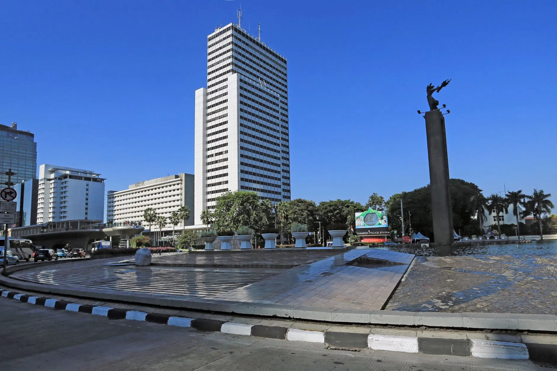 The Selamat Datang welcome monument on its fountain plaza at Bundaran HI in Jakarta, with the Thamrin high-rise skyline behind it under a deep blue sky.