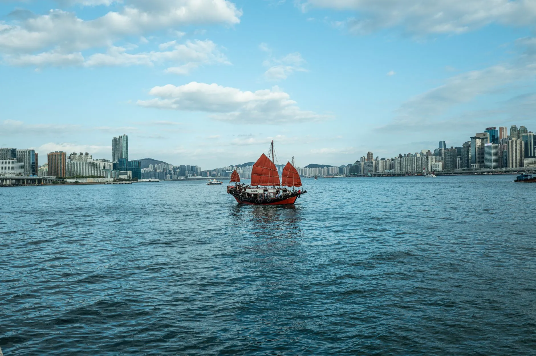 A traditional red-sailed junk boat crossing Victoria Harbour in Hong Kong, with the skylines and green hills of both shores behind it under a blue sky.