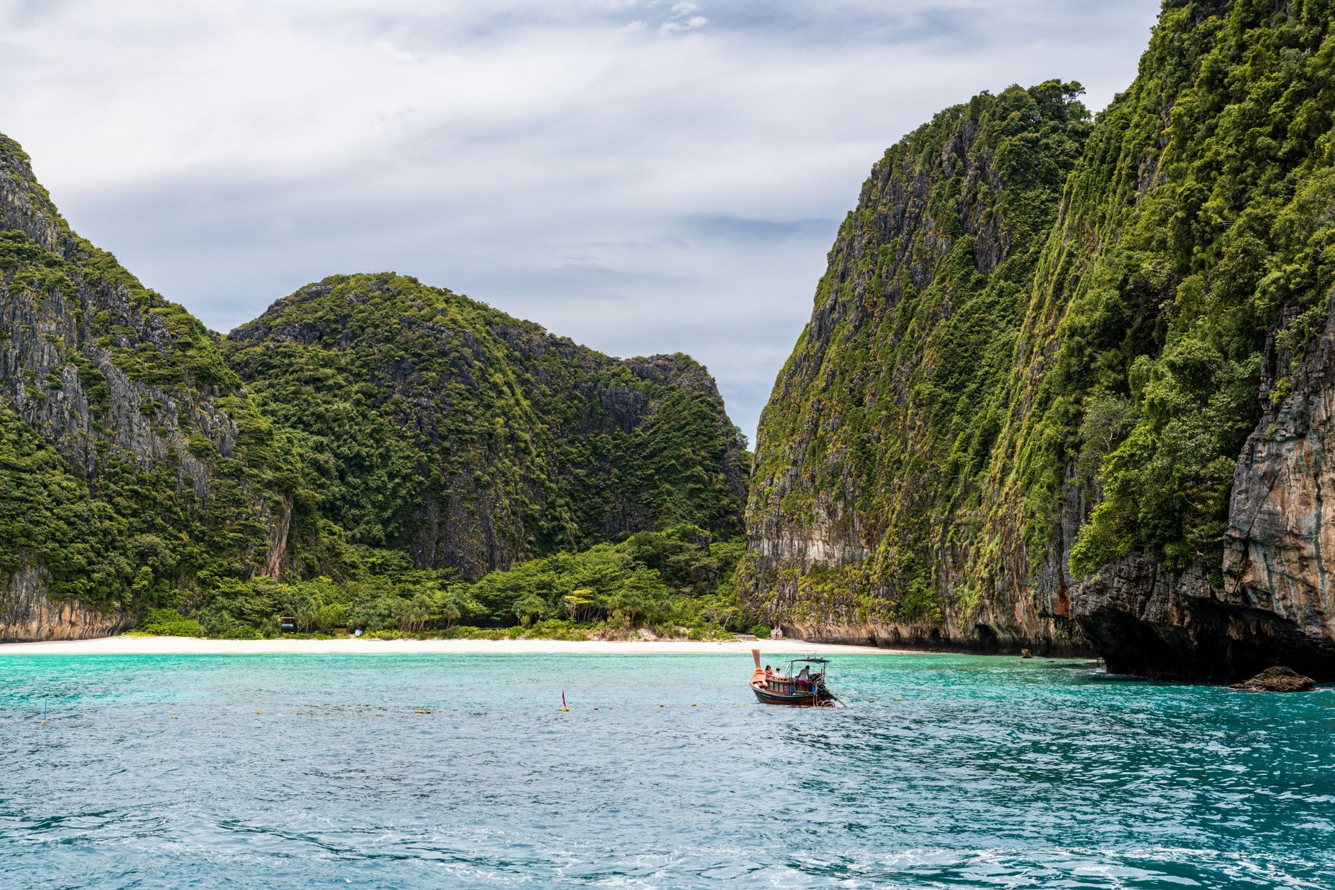 Phuket — limestone karst cliffs above a white-sand beach with a traditional longtail boat on turquoise water, Thailand