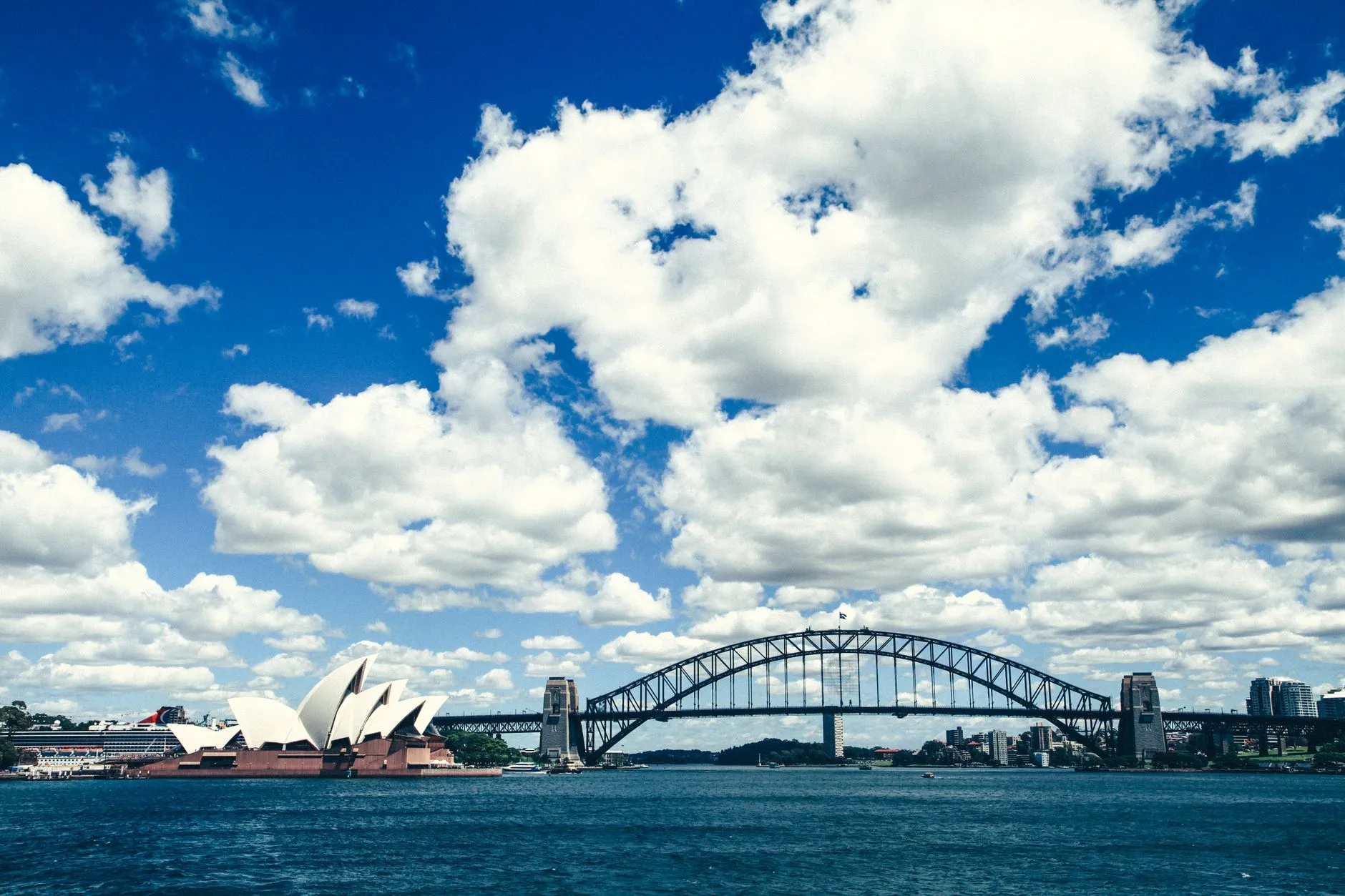 Sydney Opera House and the Sydney Harbour Bridge across the blue water of Sydney Harbour under a bright sky with white clouds.