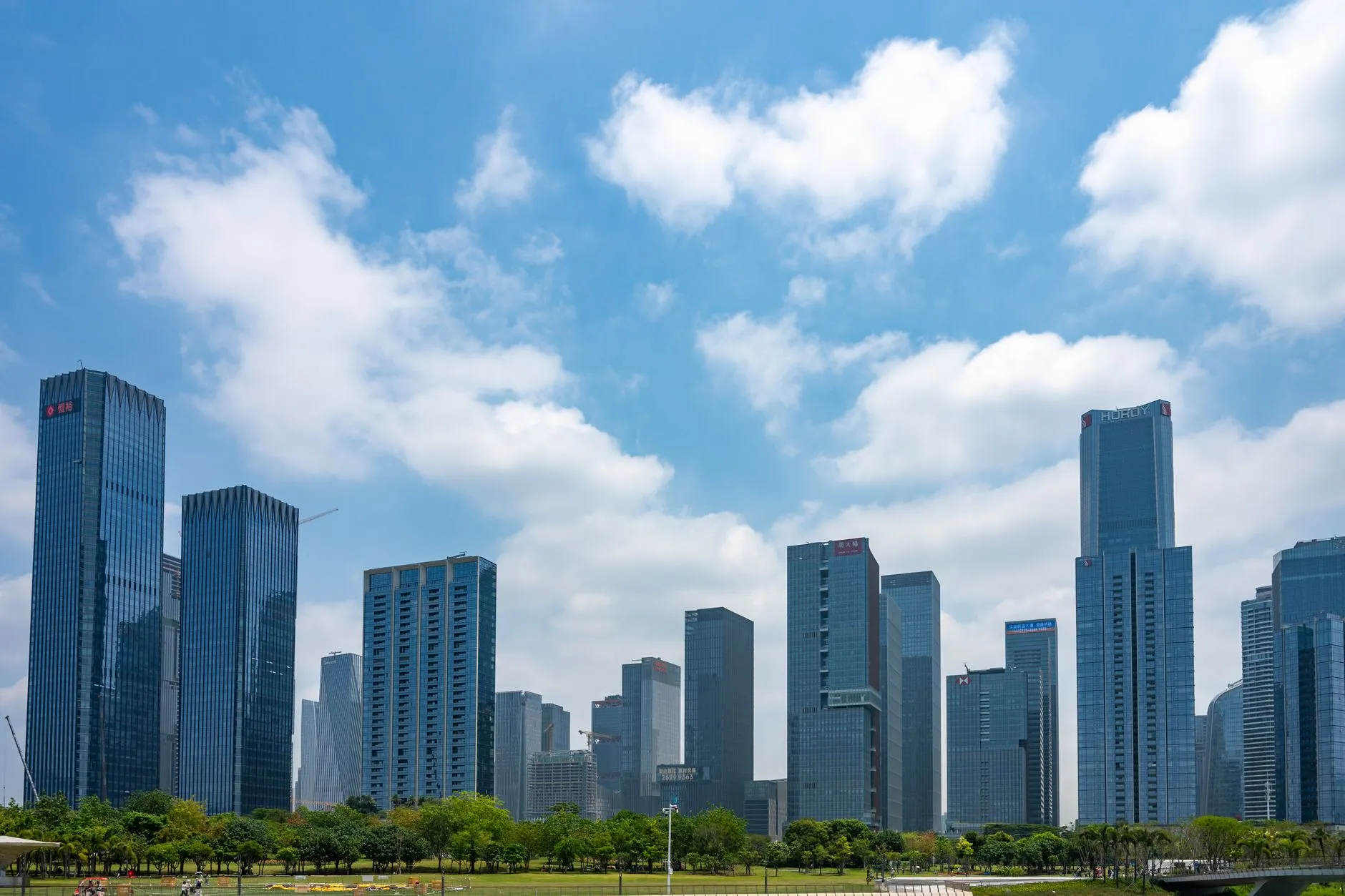 The Shenzhen skyline on a bright day, modern glass towers rising behind a green park of trees and lawn under a blue sky with white clouds.